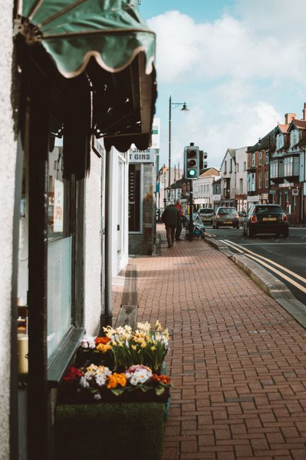 An outdoor scene of a sidewalk along Mile End Road with a flower shop from Cambridge Heath Flowers, featuring a display of fresh, colorful flowers including yellow, white, and pink blooms in a dark gr