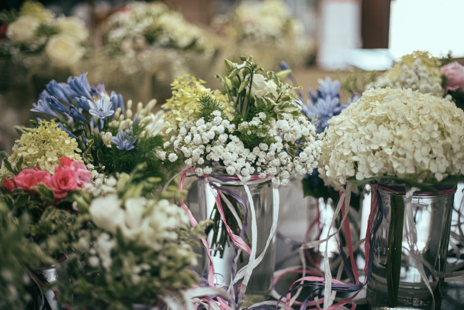 A close-up view of various floral arrangements in glass vases, featuring a mix of white hydrangeas, delicate blue and purple flowers, soft pink roses, and small white baby's breath, all arranged with 