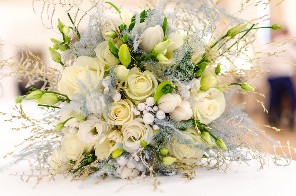 A floral bouquet arrangement featuring predominantly white roses and white tulips, accented with delicate white berries and blue-tinted foliage, arranged in a rounded style. The flowers are fresh, wit