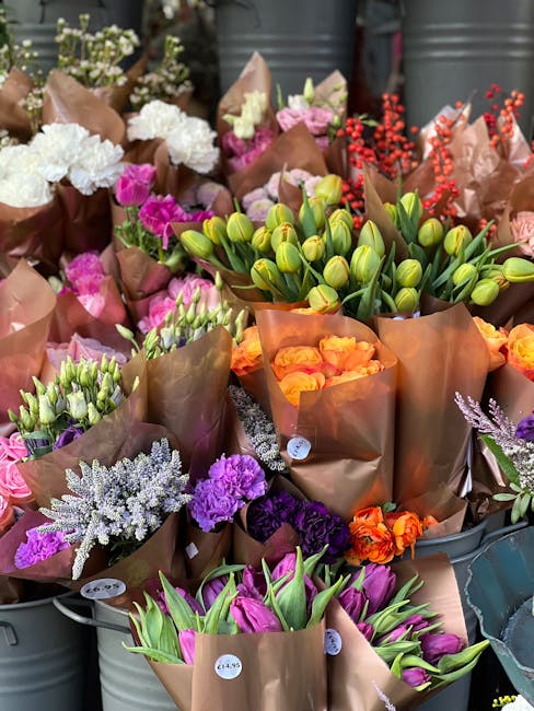 A diverse display of fresh flowers arranged in brown paper wrapping, including white and purple carnations, pink and purple roses, orange and yellow tulips, clusters of green tulip buds, and sprigs of