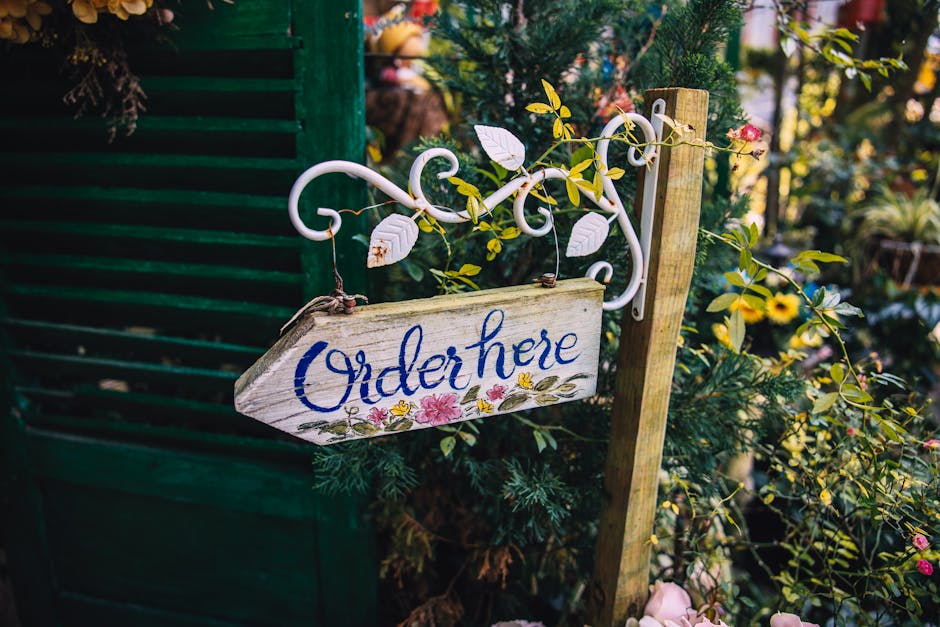 A rustic wooden garden sign with the words 'Order here' painted in blue, hanging from a white wrought iron bracket adorned with delicate scrollwork. The sign is surrounded by lush greenery, including 