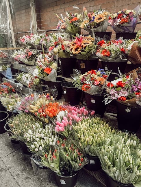 An outdoor display of fresh flower bouquets arranged in black buckets at Cambridge Heath Flowers, viewed from the front with a variety of tulips, roses, and mixed seasonal flowers in shades of pink, o