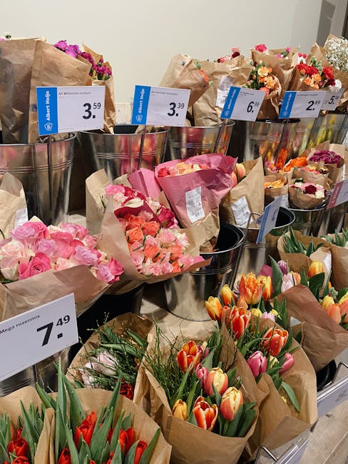 A display of various flower bouquets in a retail setting, with arrangements featuring pink roses and coral and yellow tulips wrapped in brown paper or placed in black containers. The bouquets are arra